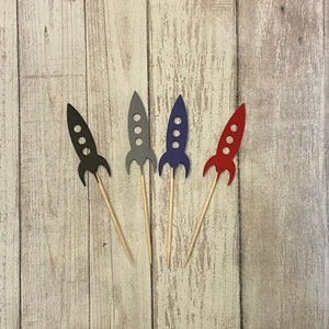 May include: Four colorful rocket ship cupcake toppers. Each rocket is a different color: black, gray, blue, and red. The rockets have three circular windows and are attached to wooden skewers. The toppers are arranged on a light wood grain surface.