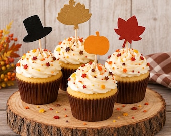 May include: Four cupcakes with white frosting and autumn-themed sprinkles, each topped with a decorative paper topper: a black top hat, a gold turkey, an orange pumpkin, and a red maple leaf, all arranged on a round wooden slice.