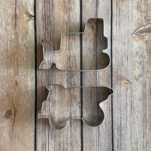 May include: Two metal cookie cutters shaped like construction vehicles. The top cutter is a dump truck and the bottom cutter is a bulldozer.