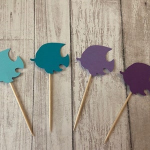 May include: Four paper fish cupcake toppers. Two are teal blue and two are purple. The fish are attached to wooden skewers.