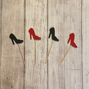 May include: Four wooden skewers with red and black high heel shoe cutouts attached to the top. The shoes are on a white wood background.