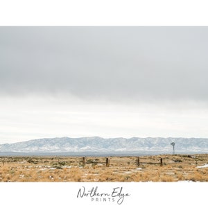 May include: A scenic landscape photograph featuring a distant mountain range with snow-capped peaks, a brown field with a wooden fence, and a single windmill in the distance. The sky is overcast and gray.