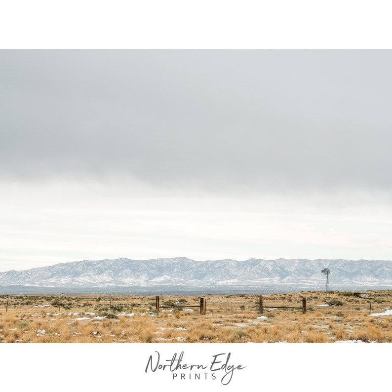 May include: A scenic landscape photograph featuring a distant mountain range with snow-capped peaks, a brown field with a wooden fence, and a single windmill in the distance. The sky is overcast and gray.