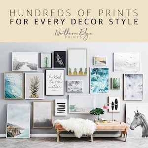 May include: A gallery wall with a variety of framed prints, including a pineapple, a wave, a cactus, a feather, a palm tree, a beach scene, and a quote that says "Be kind to one another". The wall is painted a light gray and the frames are all white. There is a pink bench in front of the wall with a white fur throw on top.