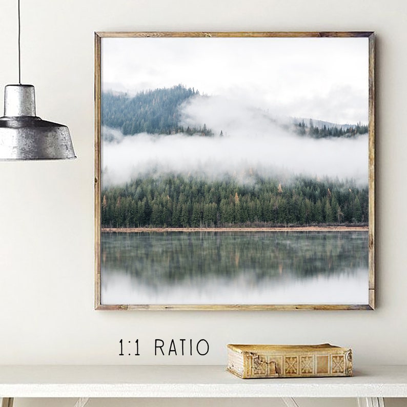 May include: A framed landscape print featuring a serene forest scene reflected in a calm lake. The image showcases a misty mountain range with evergreen trees, creating a tranquil and natural aesthetic. The frame is made of wood.