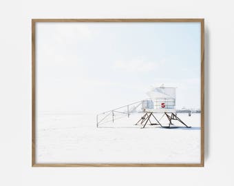 Puede incluir: Un puesto de salvavidas blanco con el número "5" en rojo en el lateral, situado en una playa de arena. El puesto tiene una escalera de metal que lleva a la plataforma. El fondo es un cielo azul brillante.