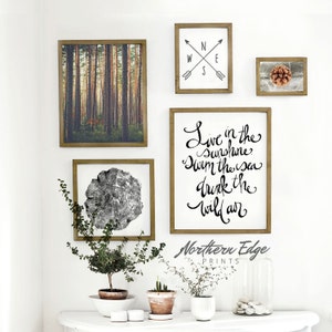 May include: A set of four framed prints with a nature theme. The prints feature a forest scene, a tree stump, a compass, and a quote that reads "Live in the Sunshine, Swim the Sea, Drink the Wild Air". The prints are arranged on a white wall above a white table with three potted plants.