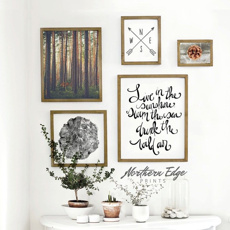 May include: A set of four framed prints with a nature theme. The prints feature a forest scene, a tree stump, a compass, and a quote that reads "Live in the Sunshine, Swim the Sea, Drink the Wild Air". The prints are arranged on a white wall above a white table with three potted plants.