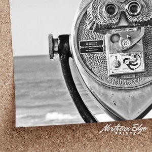 May include: A black and white photo of a vintage coin-operated viewer with the text "Quarters Only" and "To Operate" on the side. The viewer is pointed towards a body of water.