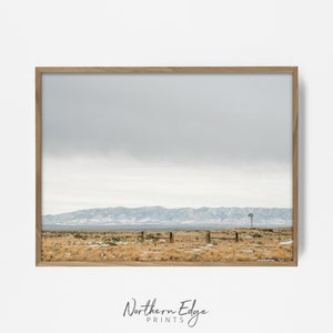 May include: A framed photograph of a distant mountain range with a single windmill in the foreground. The sky is a light gray with a few clouds. The ground is covered in dry grass and snow.