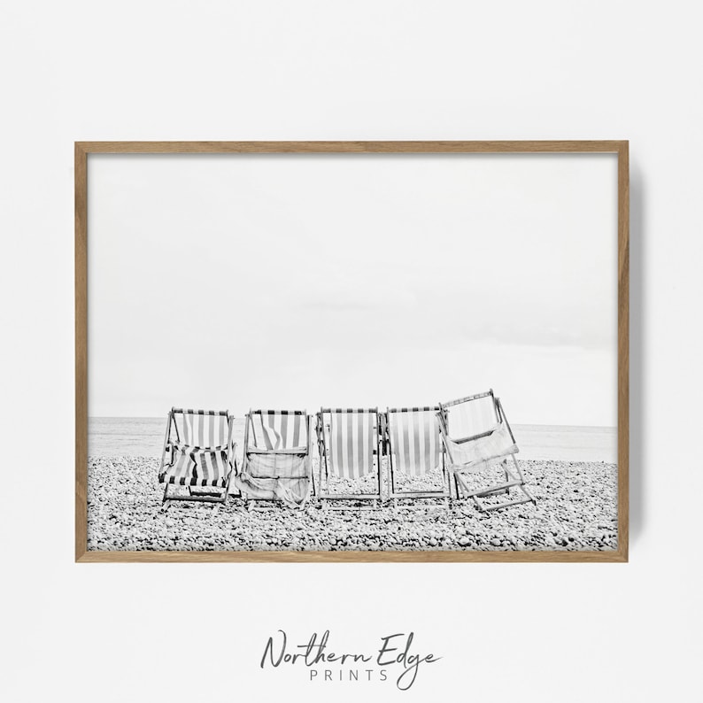 Puede incluir: Fotograf&iacute;a en blanco y negro de cinco sillas de playa a rayas en una playa de guijarros. Las sillas est&aacute;n orientadas hacia el oc&eacute;ano y est&aacute;n parcialmente plegadas. El cielo est&aacute; nublado y la playa est&aacute; vac&iacute;a.