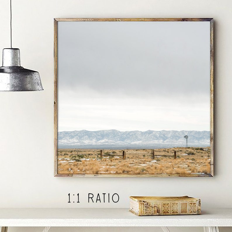 May include: A framed photograph of a desert landscape with a mountain range in the background. The sky is a light blue with some clouds. There is a fence in the foreground and a windmill in the distance.