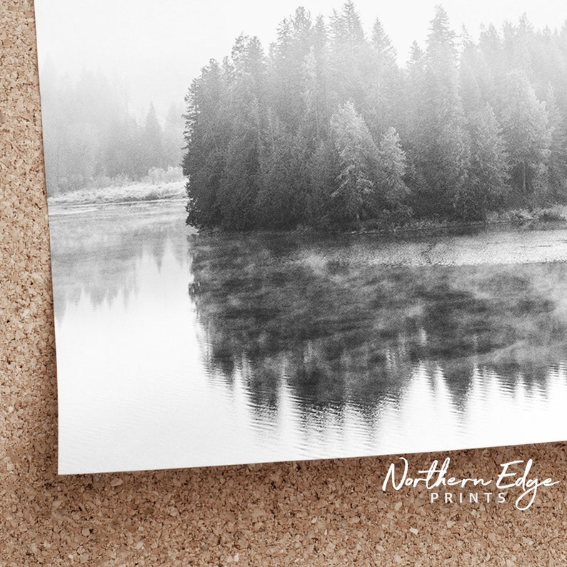 May include: Black and white landscape print featuring a misty lake reflecting a forest of evergreen trees. The print has a soft, diffused quality, with the trees and their reflection creating a symmetrical pattern. The print is on white paper.