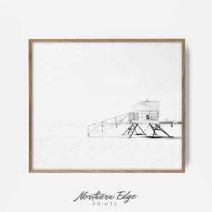 May include: Black and white photograph of a lifeguard stand on a beach. The stand is a simple structure with a ladder leading up to the top. The beach is empty and the sky is cloudy.