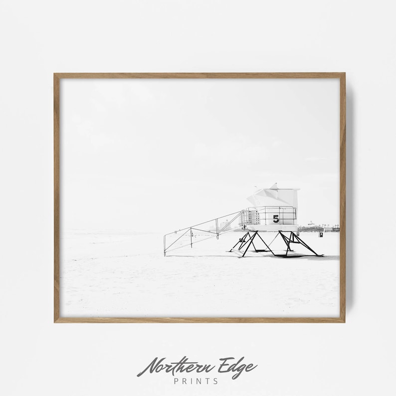 May include: Black and white photograph of a lifeguard stand on a beach. The stand is a simple structure with a ladder leading up to the top. The beach is empty and the sky is cloudy.