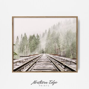 May include: A framed print featuring a railway track receding into a misty forest. The tracks are centered, leading the eye towards a backdrop of tall, green trees shrouded in fog. The print is in a wooden frame, with the text "Northern Edge PRINTS" at the bottom.