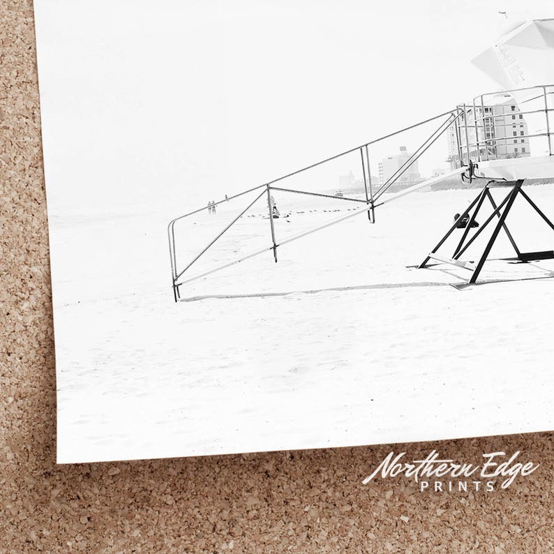 May include: Black and white photograph of a lifeguard stand on a beach. The stand is made of metal and has a ladder leading up to the top. The beach is empty and the sky is overcast.