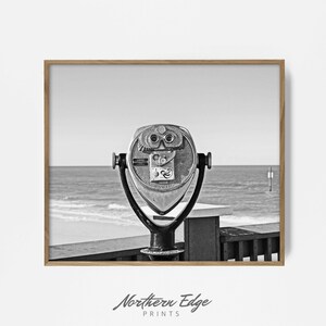 Puede incluir: Una fotografía en blanco y negro de un telescopio vintage de monedas en un paseo marítimo de madera con vistas a una playa. El telescopio está orientado hacia el océano con olas rompiendo en el fondo.