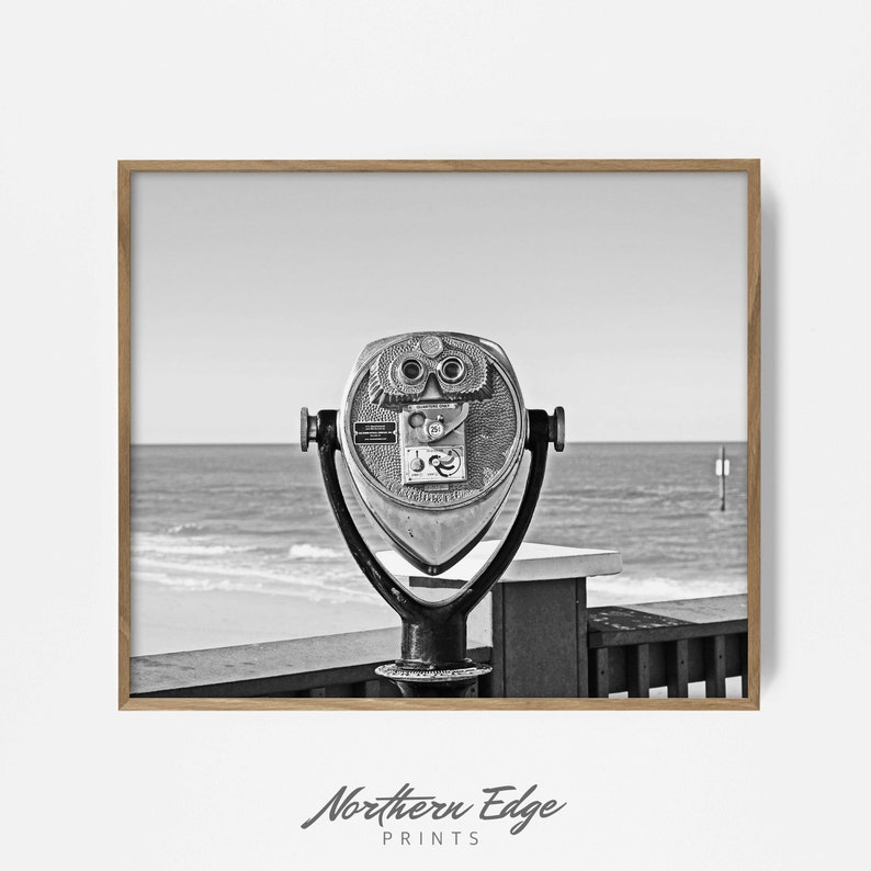 May include: A black and white photograph of a vintage coin-operated telescope on a wooden boardwalk overlooking a beach. The telescope is facing the ocean with waves crashing in the background.