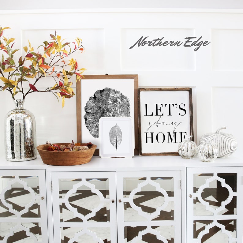 May include: A white cabinet with decorative mirrored doors displays fall-themed decor. Items include framed art, a wooden bowl with dried items, silver pumpkins, and a mercury glass vase with fall foliage. The text "LET'S stay HOME" is visible.