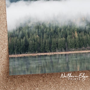 May include: A landscape print featuring a tranquil lake reflecting a forest of evergreen trees. The upper portion of the image is shrouded in a layer of mist, creating a serene and atmospheric scene. The print is labeled "Northern Edge Prints".