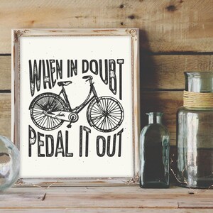 K&ouml;nnte beinhalten: Schwarz-wei&szlig;-Druck mit einem Fahrrad und dem Text "When in doubt, pedal it out". Der Druck ist in einem wei&szlig;en Rahmen gerahmt und liegt auf einer Holzoberfl&auml;che.