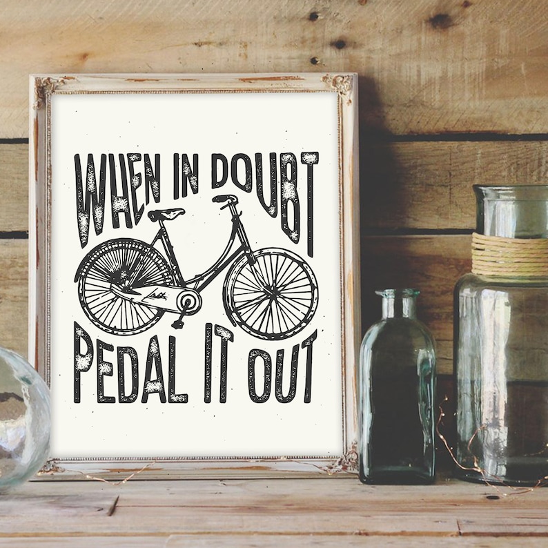 K&ouml;nnte beinhalten: Schwarz-wei&szlig;-Druck mit einem Fahrrad und dem Text "When in doubt, pedal it out". Der Druck ist in einem wei&szlig;en Rahmen gerahmt und liegt auf einer Holzoberfl&auml;che.
