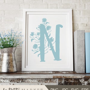 May include: A white frame with a light blue letter N surrounded by a floral design. The letter N is a silhouette with a white background. The floral design is a silhouette with a light blue background. The frame is sitting on a wooden surface.
