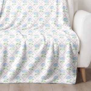 May include: A white throw blanket with a repeating pattern of blue and green hydrangeas on a white background with pink diamond shapes.