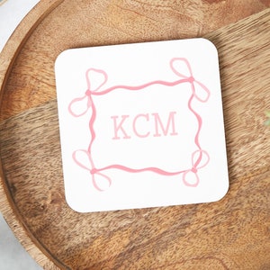 May include: White coaster with a pink ribbon border and the initials "KCM" in the center.
