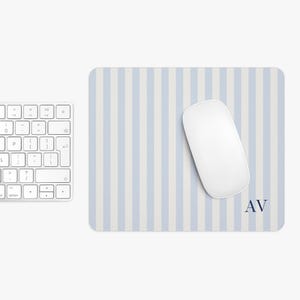 May include: A white wireless mouse rests on a blue and white striped mousepad. The mousepad has the initials "AV" in navy blue in the lower right corner. A white keyboard is partially visible on the left.