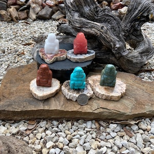 May include: Six small Buddha figurines in various colors, including white, red, brown, turquoise, and green. Each Buddha sits on a small, round, polished stone base. The figurines are displayed on a larger stone surface.