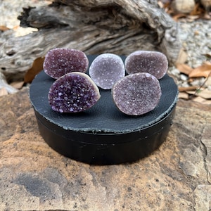 Peut inclure: Cinq géodes d'améthyste de différentes nuances de violet et de lavande sont exposées sur une plateforme circulaire noire. Les géodes ont une structure cristalline et sont disposées sur une surface noire.