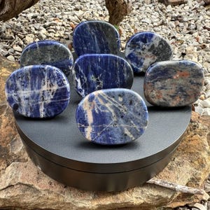 May include: A selection of oval sodalite stones, each displaying a rich blue hue with white and grey veining. The stones are presented on a black circular platform, highlighting their natural patterns. The stones' sizes and colours vary, offering a unique aesthetic.
