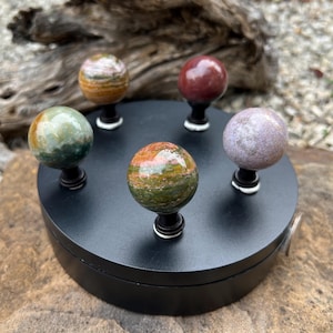 Op de afbeelding: Een zwarte displaystandaard houdt vijf gepolijste edelsteenbollen in tinten groen, rood, oranje en paars. Elke bol is gemonteerd op een zwarte steel met een witte accent. De bollen zijn in een cirkelvormig patroon gerangschikt.