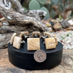 May include: A collection of square-shaped bottle stoppers with a light brown, sandstone-like appearance. The stoppers have a dark metal base and are arranged on a black circular stand. A U.S. quarter is included for size reference.