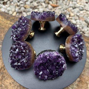 May include: A white ceramic mug with a black handle and the text "But First, Coffee" in a black, bold font. The mug is on a wooden surface. The mug is approximately 10 cm tall.