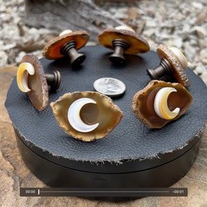 May include: Six agate and mother of pearl cabinet knobs with a crescent moon design. The knobs are displayed on a black circular box with a brown leather-like surface.