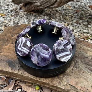 May include: A selection of amethyst gemstone cabinet knobs and pulls with gold-coloured hardware. The knobs are a mix of round and rectangular shapes, displaying the natural purple and white banding of the stone. The knobs are displayed on a black rotating platform.