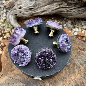 May include: A selection of amethyst crystal cabinet knobs with gold-coloured hardware. The knobs are arranged on a black leather-like surface. The amethyst crystals display shades of purple and are in various shapes.
