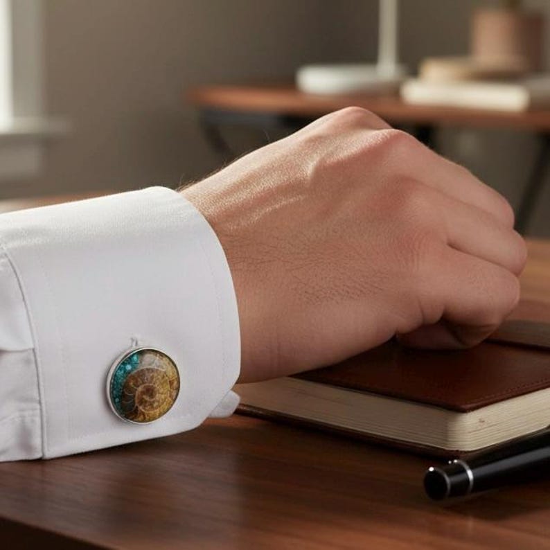 Pu&ograve; includere: Primo piano di un polsino di camicia bianca con un gemello d'argento. Il gemello presenta un design rotondo con un motivo di ammonite fossilizzata nei toni del marrone, verde acqua e oro. Il gemello &egrave; posto su una superficie di legno scuro.