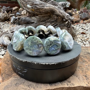 May include: A collection of circular cabinet knobs featuring polished moss agate stone faces with intricate green and white dendritic patterns, each mounted on a dark bronze-toned metal base, arranged in a cluster on a black display stand.