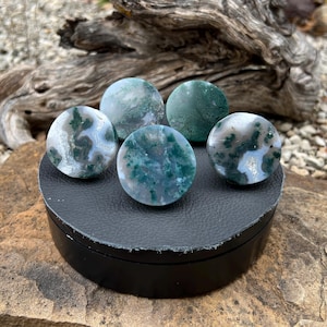 Pode incluir: Cinco pedras redondas e polidas com um padrão de ágata musgo em tons de verde, branco e cinza. As pedras são exibidas em uma plataforma circular preta. O fundo apresenta um pedaço de madeira flutuante e um ambiente natural ao ar livre.