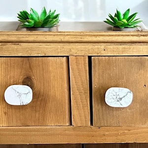 May include: Two wooden drawers with white marble-patterned knobs. The drawers are on a wooden cabinet with a light brown finish. Two small green succulent plants are on top of the cabinet.