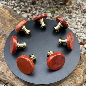 Może przedstawiać: Zestaw siedmiu czerwonych gałek do szafek z jaspisu ze złotymi metalowymi podstawami, ułożonych w okrąg na czarnej powierzchni. Gałki mają gładki, zaokrąglony kształt i naturalny, ziemisty wygląd.