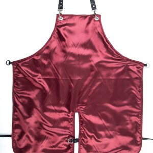 May include: A burgundy satin apron with black leather straps and silver buckles. The apron has a split in the front and is designed for use in a salon or barbershop.