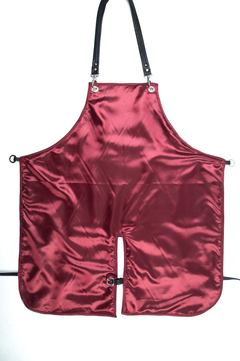 May include: A burgundy satin apron with black leather straps and silver buckles. The apron has a split in the front and is designed for use in a salon or barbershop.