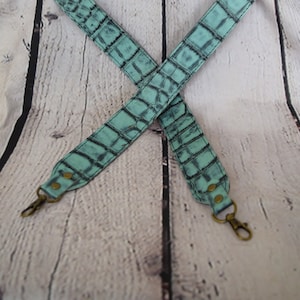 Peut inclure: Une bandoulière d'appareil photo en cuir bleu clair avec un motif crocodile et des ferrures dorées. La bandoulière est croisée et posée à plat sur un fond de briques blanches.