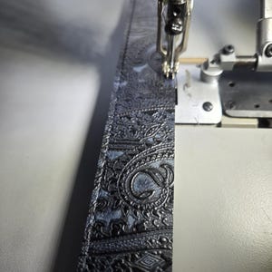 May include: A close-up shot of a sewing machine in operation, stitching a black paisley fabric. The fabric has a detailed embossed pattern in dark grey. The machine's needle is visible, creating a seam along the edge of the material.