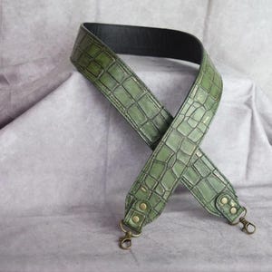 Op de afbeelding: Een brede tasriem met een textuur van olijfgroen krokodillenleer aan de buitenkant en een gladde zwarte binnenkant. De riem is voorzien van metalen messingkleurige hardware met draaibare karabijnhaken aan beide uiteinden voor bevestiging.
